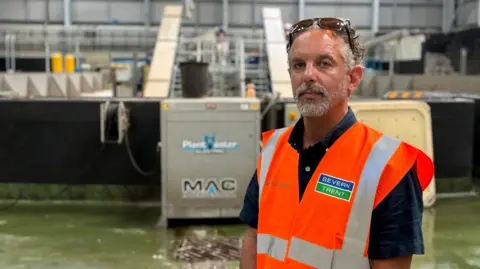 Chris Lofts has curly grey hair and a short grey beard. He wears a dark blue short-sleeved shirt underneath a bright orange hi-vis vest with Severn Trent's logo emblazoned on the front. 