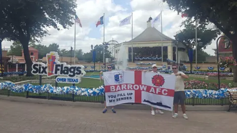 Three men hold up a large England flag with the words Barrow AFC across the horizontal line of the red cross. In the top left corner it has the Barrow AFC badge, the top right has a red rose, the bottom left has the phrase "you fill up my senses" and the remaining quarter has the outline of a black submarine. The three men are all wearing sunglasses and are standing in front of a sign for the theme park Six Flags Over Texas. There is a pavilion and a rollercoaster in the background.