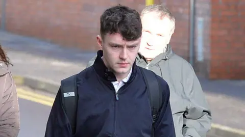 A man in his early 20s with dark hair and wearing a navy blue jacket walking into court. He is looking down slightly and scowling.