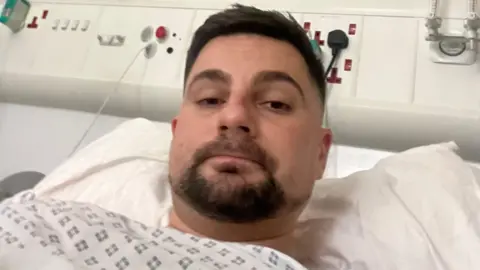 Joe Harris with a swollen neck while at Gloucestershire Royal Hospital for treatment. He is laying on a hospital bed and is pictured from the neck up. He is looking directly at the camera and is unsmiling.