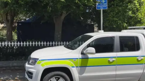 BBC A white police vehicle with a yellow stripe outlined in blue. The vehicle is parked and has dark windows. There is a black and white fence behind with trees behind.
