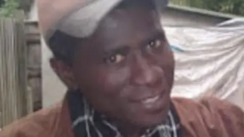 A blurry photo of a man wearing a baseball cap and a brown jacket, smiling at the camera.