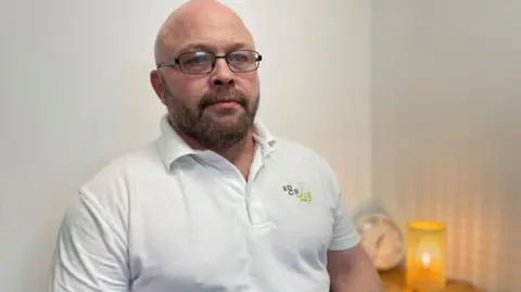 A man with shaved hair, a beard and glasses sits in a white polo shirt. Next to him on a side table is a lamp and clock