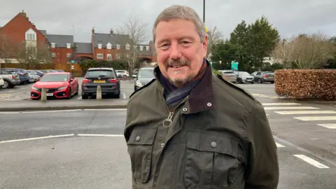 A man with grey hair, wearing a dark green jacket, smiles at the camera with hsi arms behind his back. A car park is behind him.