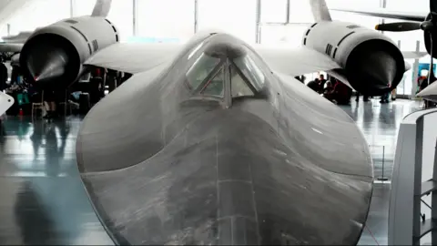 John Fairhall/BBC A shot of a SR-71 Blackbird in a hangar at IWM Duxford. The black plane is seen from above and it has a flat body, a bit like a manta ray. Its nose has been cut off in the image. Beyond the body of the plane are two wings with very large engines. 