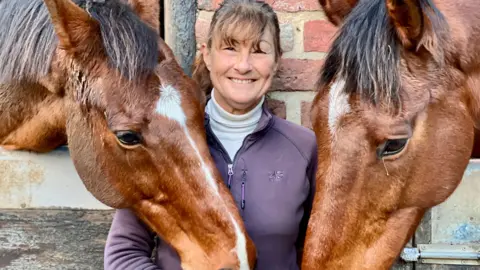 BBC/Linsey Smith Melanie Watson, wearing a purple tracksuit top and white polo neck stands in between two brown horses.