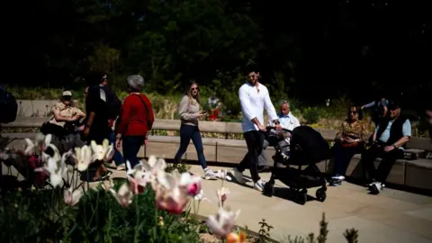 PA People of a range of ages enjoy the park, with pink and white tulips in the foreground of the image. People sit on wooden benches on round pathway. 