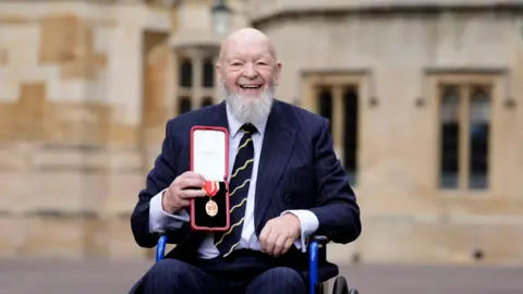 PA Media Michael Eavis is in a wheelchair in front of Windsor Castle. He is holding up a golden medal. He is wearing a blue suit and is laughing into the camera.