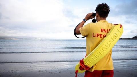 Lifeguard with red shorts and yellow top with yellow rescue tube across his back looking out to the sea off Weymouth with binoculars