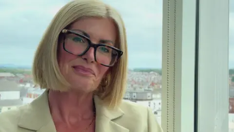 An image of Vicky Gent, she is blonde and wearing tortoiseshell glasses, and a beige blazer. She is sitting in front of a window with Blackpool in the background.