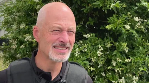 A Trading Standards officer, who is bald with a grey beard, standing in front of a shrub with white flowers.