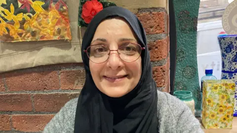 A woman with a black headscarf smiles at the camera. She is sitting in front of a brick wall and there are colourful vases to her right