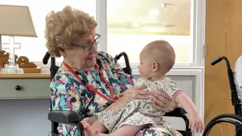 BBC An older lady in a wheelchair with brightly coloured blouse holds a six-month-old baby who is playing with her lanyard. She sits in a wheelchair as we can see the black handles behind her head and the armrests to the sides.