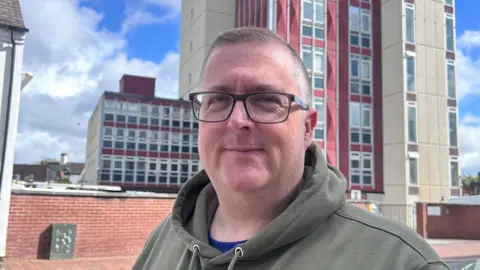 Ben Dolan stands in front of the BT building in Stafford. The building is a large, square tower block, with red and cream panels. Ben has short grey hair, black rectangle framed glasse and is wearing a green hoody with a blue tshirt visible underneath. 