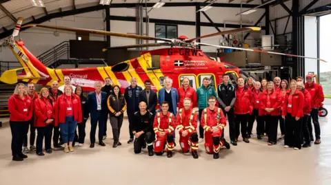 Cornwall Air Ambulance Cornwall Air Ambulance staff stand and kneel in front of a helicopter flanked by Formula One representatives in a large hangar