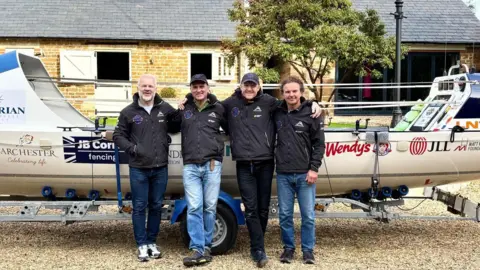 Ollie Phillips is standing far left wearing jeans, trainers and a black waterproof jacket with three other friends. An ocean rowing boat is in the background.