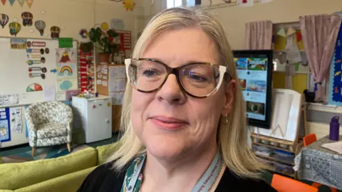 Hilary Cunningham is in a classroom, she is wearing white and black glasses and a brown top. She has blonde hair. Behind her is a white board with decorations, a chair, a white fridge and an interactive board. The cream walls are covered with displays. 