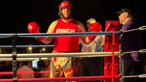 Craig Hinde Craig Hinde wearing red boxing gear and gloves while standing in a boxing ring.