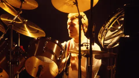 Getty Images Rick Buckler behind a drum kit on stage, against a black background. He and his drums are lit up with an orange-sepia colour.