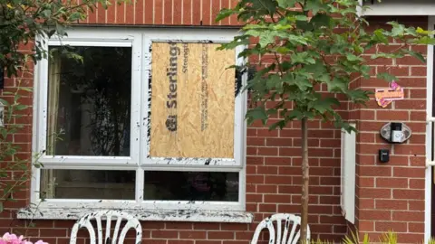 Close shot of a damaged window on a red brick building. Plywood has been put over smashed glass.