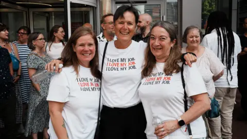 John Fairhall/BBC Three women wear white T-shirts with the words "I love my job" written in black and red writing. Other members of staff, some in white T-shirts, gather behind them outside the University of Essex's Southend campus