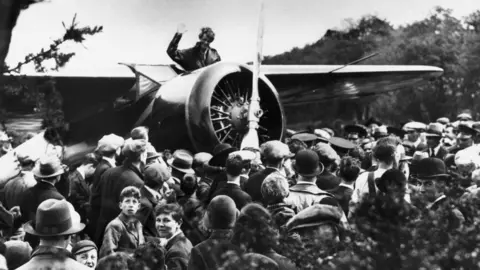 Black and white picture of lots of different people wearing hats, looking up at women leaving a plane. 
