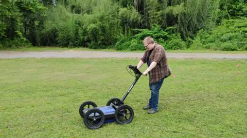 Duffy's Cut Project A man with fair hair and glasses and wearing a checked-shirt and jeans pushes a four-wheeled machine along a field of grass
