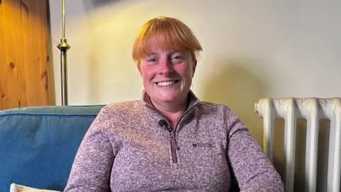 Kate Bradbrook/BBC Hannah with short ginger hair smiling at the camera while wearing a pink zip-up top. She is sitting in a blue armchair with a white wall and radiator behind.