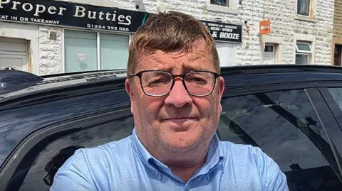 David Fielding with brown hair standing in front of a black vehicle and a sandwich shop wearing black glasses and a blue shirt.