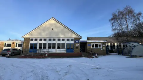 Shaun Whitmore/BBC A 1960s/70s single-storey school building with an apex roof. It has blue double doors at each side of the school hall, and a car is parked in the car park, which is covered in snow. There is a plastic bike shelter in the car park, too.