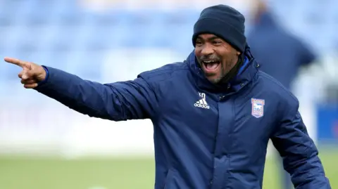 Kieron Dyer wearing a navy beanie hat and navy coat with the Ipswich Town and Adidas emblems on. He is laughing with his mouth open and standing on the pitch at a stadium, which has blue seats behind him. Dyer is pointing at something out of sight to his right. 