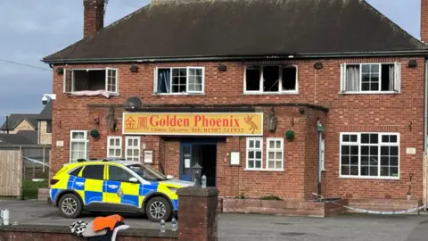 Joe Weir/BBC Shot of a fire-damaged building with a police car parked at the front. It has Georgian-style windows and a sign which says Golden Phoenix above the door.
