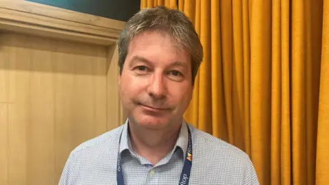 A man wearing a black and white shirt and a lanyard, is standing in front of an orange curtain.