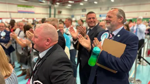 A group of men in dark suits, some wearing Reform blue rosettes, clapping in the Benham Sports Arena.