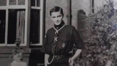 SVLB A black and white photograph of Geoffrey Crawford standing next to a shrub outside of a house. His short dark hair is combed to the side. He is wearing a dark shirt, trousers and a tie.