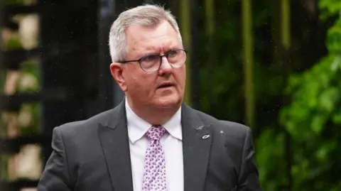 A man with round glasses and white hair looks to his left. He is wearing a dark suit, white shirt and pink patterned tie. He is wearing a small fish shaped badge on his suit.