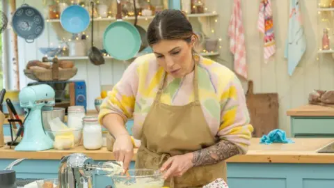 Love productions Georgie Grasso cooking in the Bake Off tent. She has a beige apron on and underneath has a pink, yellow and green jumper. She is looking down at the table and is stirring some sort of whipped dessert in a bowl.