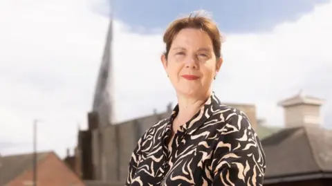 A woman with brown hair stood in front of the camera with Chesterfield's crooked spire in the background. 