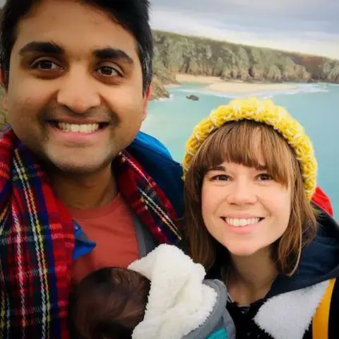 Angharad Korambayil is seen on the left, with a baby on his chest, with Anghard on the right, both smiling into the camera with a seascape behind them. 