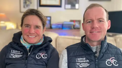 James Burridge/BBC Piggy and Tom March sitting side by side on a cream sofa, both wearing dark blue gilets with Spinal Research and British Eventing Support Trust logos on one side of the chest, and a cycling logo on the other. A table lamp, sideboard, framed pictures on the wall and a TV can be seen behind them. 