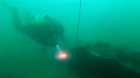 A diver with air tanks on their back shines a light, red with a white glow at the centre, as they near a long rocky looking base, the remains of a shipwreck. They are in green water, what appears to be a cable extends upwards from the wreck.