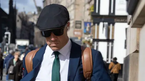 Alan Simpson A man keeps his head down as he walks along a street. He is wearing a black flat cap, sunglasses, a blue jacket, white shirt, green tie and has leather rucksack handles over his shoulders