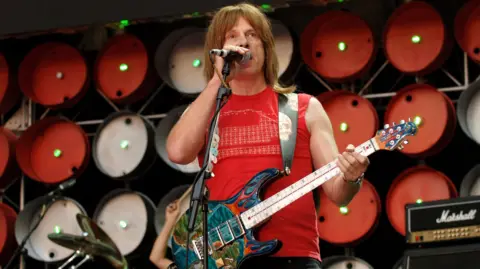 Getty Images Christopher Guest of Spinal Tap addresses the crowd through a microphone during the Live Earth concert at Wembley Stadium. He has long brown hair and is wearing a red top. The unique guitar is hung around his neck