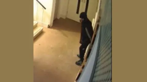 The man who they wish to speak to on CCTV. He is described as white, of slim build, with a black beard. He is wearing a beanie, a dark-coloured jacket with white stripes down the side, dark jogging bottoms and a pair of blue trainers.