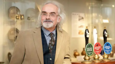 Stephanie Belton David Thorold has grey shoulder length hair and a grey moustache and beard. He wears unrimmed glasses and is looking at the camera. He wears a light brown jacket, greem-checked waistcoat, blue and white floral shirt and a solar system-themed tie. He is leaning on a bar with three beer pumps with a glass museum presentation case in the background.