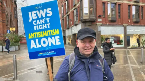 Olivia Richwald/BBC A man in a blue waterproof coat and black cap holding a placard which reads 'Jews can't fight antisemitism alone'. He is in Leeds city centre and it is raining.