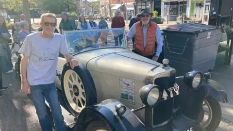 Two men standing either side of a vintage car with their hands on the sides of it. They are both wearing sunglasses. The man on the left is wearing a T-shirt and jeans and the man on the right is wearing an orange gilet and a bush hat. It is a sunny day and there are people in the background standing in a street, which has small shops on it. 