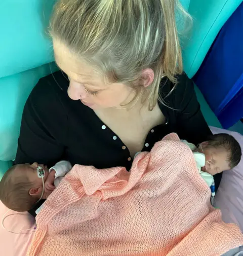 Brioney Garrett Brioney holds her newborn identical twin daughters in 2022 - they lie either side of her as she sits up in a chair, wearing a black top. The babies bodies are covered with an orange blanket.