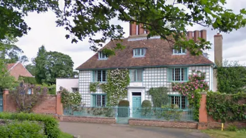 Google A three-storey village house with a pattern of blue-edged squares on a white background on the front elevation. There is blue door in a white frame. Climbing plants are evident on the facade. There is a small paved area in front of the door with shrubs. There are brick walls either side of the house covered in leaves. A low blue railing separates the property from the road.