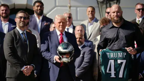 US President Donald Trump holds a helmet as he poses with Philadelphia Eagles head coach Nick Sirianni and offensive tackle Lane Johnson at an event welcoming the 2025 Super Bowl Champion Philadelphia Eagles on the South Lawn of the White House